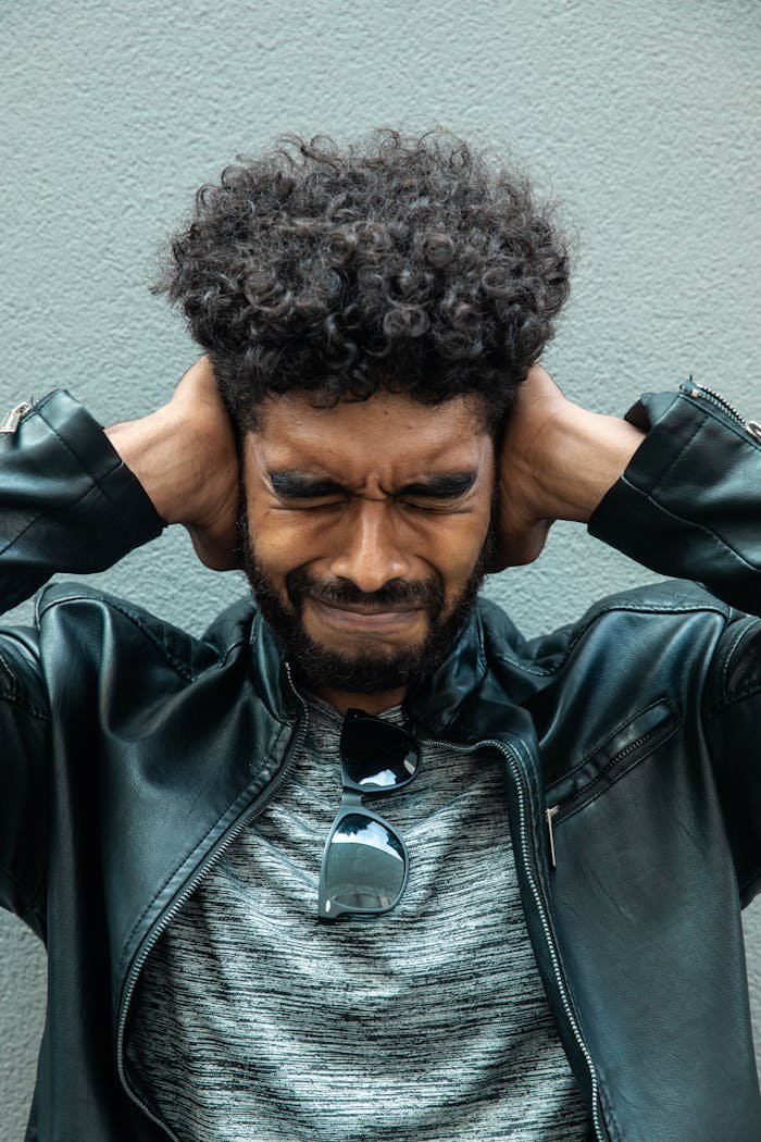 A man with curly hair and a beard shows frustration while covering his ears.